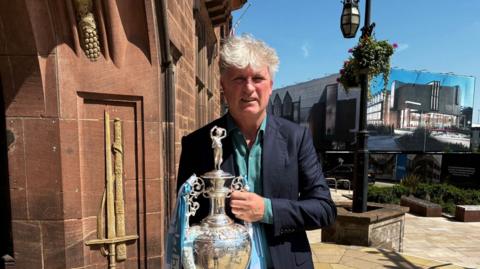 A man with grey hair holding a silver football trophy outside a council house. He has a blue jacket and green shirt on