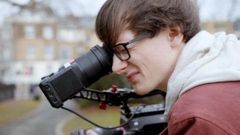 Teenage boy using a video camera. He is wearing a red jacket and a grey hoodie.