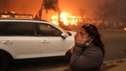 A woman wearing a facemask holds her head in her hands as behind her a wildfire rips through buildings. There's an orange glow in the sky.