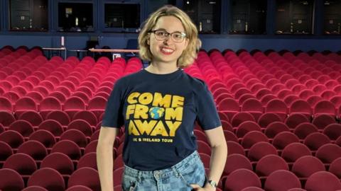 Lydia Greatrix is stood in a theatre, wearing glasses and a Come From Away tour T-shirt, with rows of red theatre chairs behind her.