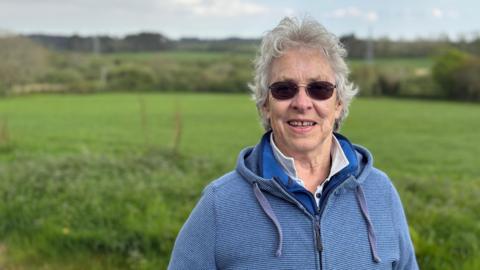Kate has curly grey hair and round sunglasses. She wears a blue hooded top and stands in front of the fields where the solar farm will go. 