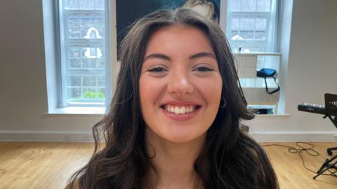 A young woman with long brown hair, smiling at the camera. She is standing in a room with a white wall and two long windows in the background. 