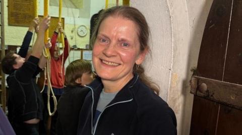 A woman with long, blonde hair smiling at the camera. People are pulling on ropes from the ceiling over her shoulder.