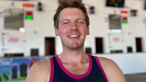 A man smiles into the camera. He is wearing a navy and pink vest and has short brown hair. He is standing in front of a sheep shearing stage.