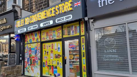 Otley Road Convenience Store. The shop has yellow posters in the windows with pictures of food and drink items.