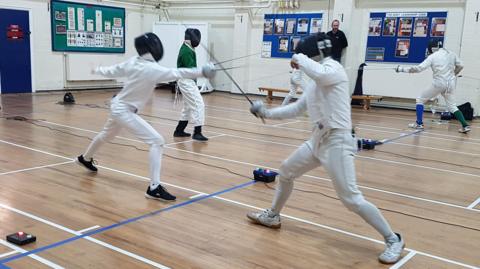 Four fencers compete wearing masks and white protective clothing in a hall with a wooden floor and lines marked out.  A coach watches over them.