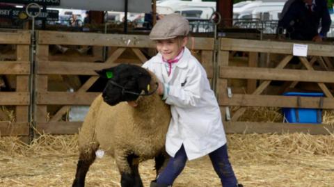 A girl holds and stands next to a sheep within a pen at the Suffolk Show that is filled with straw. She wears a white coat, flat cap and blue jeans with boots. The sheep has white wool and a black face and legs.