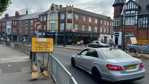A silver mercedes driving in a town centre with a yellow road closed sign to the left of the carriageway.