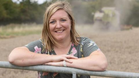 Milly leans on a metal farming gate and smiles at the camera. She has shoulder length blonde hair and wears a grey t-shirt with pink and yellow flowers. In the background, blurred, is a combine harvester and brown fiel.