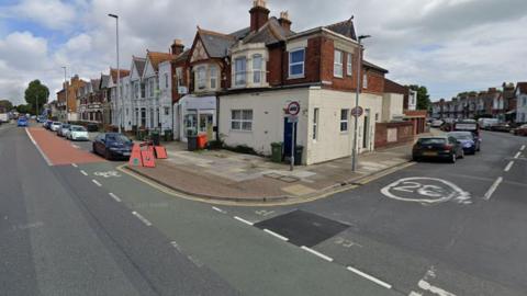 The London Road junction with Hewitt Road in Portsmouth on a cloudy day. There's no one on the street, a few cars are parked by the rows of houses.