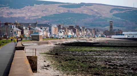A seafront view of houses and a beach, covered in mud and seaweed. Hills are visible in the background.