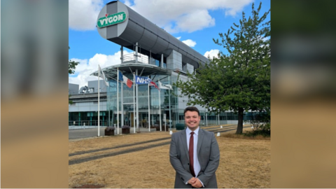 Councillor Jake Chandler standing in front of a futuristic looking glass-panelled building with flags in the foreground which is the former Motorola building (now Vygon UK) in Swindon.