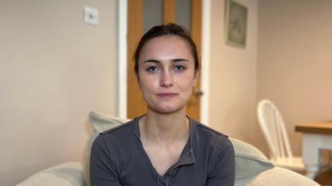 Nicole Sheppard pictured sitting in her house. She is wearing a grey top and has dark hair tied back. She is looking at the camera with a disappointed expression.