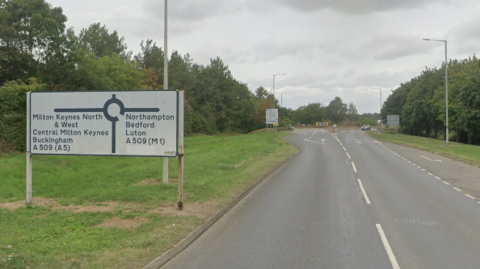 A generic view of Tongwell Street in Milton Keynes approaching a roundabout. A white road sign detailing routes off the roundabout can be seen sat on the left hand grass verge.