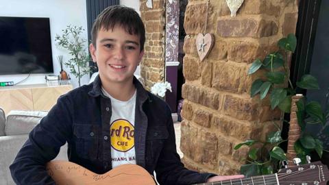 Jakob Wojcik, who has short black hair and is wearing a black open shirt over a white T-shirt. He is holding a guitar, which is signed along its top body edge, and is smiling. He is standing a TV behind him on the wall and a brick wall to his right. 