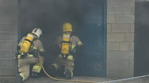 Two firefighters with a yellow hose are outside of blue doors. They are surrounding by black smoke 