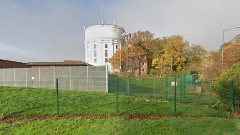 A round, white water tower set in grassland but surrounded by other water works buildings, trees and green metal security fencing.