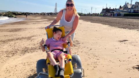 A woman wearing a vest and shorts and sunglasses positioned behind a yellow beach wheelchair with big grey wheels. In it is a young girl wearing a red-and-white school dress. They are on a wide expanse of sand on a seafront.