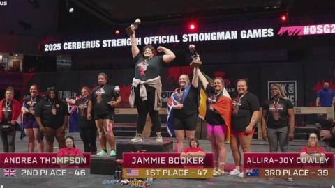 A group of athletes are stood on and around a podium after the world's strongest woman competition. Jammie Booker is on the central podium holding up a trophy.
