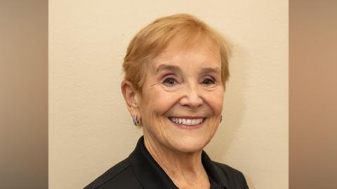 An elderly woman is smiling. She has short-cropped strawberry blonde hair and is wearing earings and eyeshadow. 