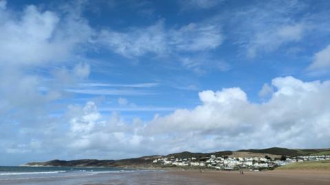 A partially cloudy sky above low hills and a coastal town, with a wide beach in the foreground