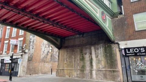 A light green bridge with a red underneath is pictured running across a street with shop fronts either side.