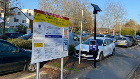 Station Road Car Park. The car park has a white and yellow sign with instructions written on it. The sign has a black stripe across it, which says "PLEASE PAY HERE" in white text. There is a solar panel powered pay and display machine with many cars parked in their bays. 