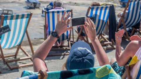 A woman sits in a striped deck chair on a beach in Cornwall. She is holding up her mobile phone to take a picture. There are more deck chairs in the background, along with small boats sitting on the sand. 