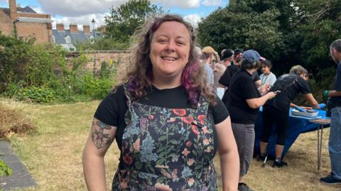 Programme organiser Lizzy standing in front of the gardening group working on their project. She is wearing floral dungarees with a black T-shirt underneath. Her hair is brown with purple streaks and she has a septum piercing and a tattoo on her left upper arm.