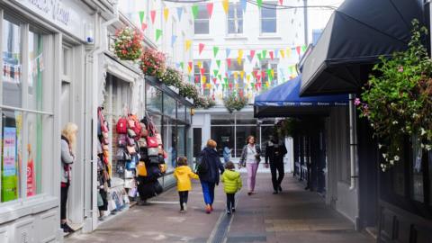 A paved street in St Peter Port lined with shops. There is a woman holding the hands of two children walking down it and a man and a woman walking up it. A lady is standing in a shop door way. There is mulit-coloured bunting strung across the lane and one of the shops has bags hung outside it.