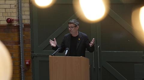 Burnham speaking at lectern during event, has both his hands raised