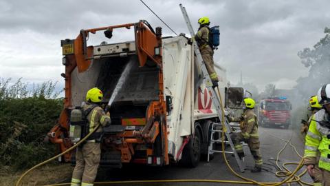 Fire fighters work on a white bin lorry, with one spraying water into the rear and another on a ladder inspecting the top.