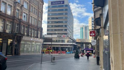 A Premier Inn hotel with a HSBC bank below. In front of the building are barriers and a fire engine. There are also shops and other businesses in view. 