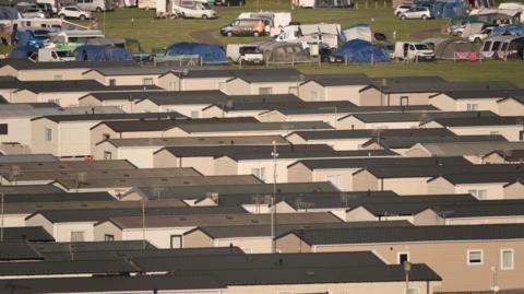 The roofs of multiple rows of static caravans stretch out across a field. Beyond the caravans, there are tents and parked vehicles dotted around the site.