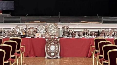 A table of trophies, with a large shield in the foreground and chairs on either side, there is a stage in the background.