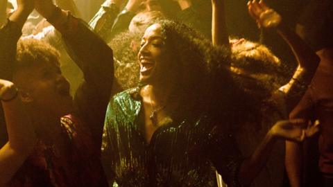 A woman wearing a glittery green top is dancing in a night club. She is smiling and surrounded by people