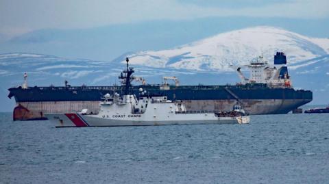 The tanker is a large ship with rusty marks on its hull. The upper part of its hull is painted blue and it has a white bridge and a single blue funnel. In the foreground is a white US Coast Guard vessel. There are snow-covered hills in the background.