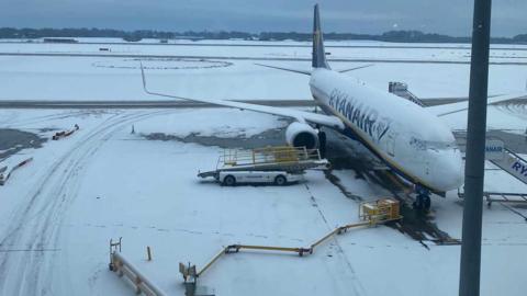 A Ryanair plane covered in snow on a Manchester Airport runway, close to baggage vehicles and equipment. The picture is taken from a terminal building.