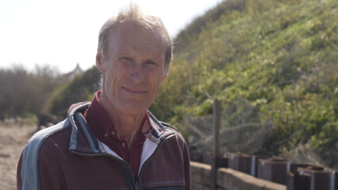 James Crowhurst standing on the beach at Thorpeness. He has short light brown hair and is squinting slightly in the sun, wearing a maroon fleece over a maroon shirt. Behind him is a wooden barrier and behind that sheet metal piling, installed in a zig zag pattern. Rising up from it is a shallow leafy cliff. 
