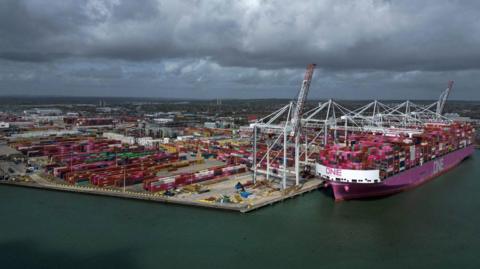 An aerial short of the port of Southampton. A row of cranes can be seen along the waterside, hanging over a large cargo ship with piles of containers.