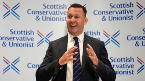 Russell Findlay gestures with his hands at the media conference for his election as leader. He is standing in front of Scottish Conservative party branding.