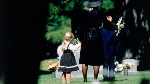 A girl and boy stood near a woman all wearing dark clothing. They are stood near a grave on a sunny day.