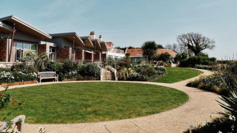 The hospice has units which are single storey and have balconies. There is circle of lawn with a wooden seat overlooking it and a path around it. There are also flower beds and palm trees.