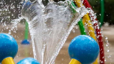 A spout of water heading upwards with droplets falling off either side. There are several colourful items blurred in the background.