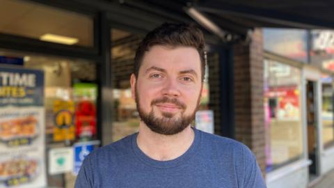 A man with dark hair and a short beard stood in front of a fish and chip shop. He is wearing a blue top.