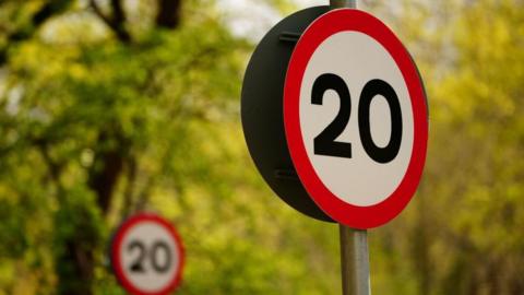 Two 20mph signs against a leafy backdrop