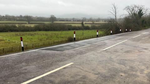 A resurfaced country road, with white markings on and reflective bollards to the side of it, surrounded by fields