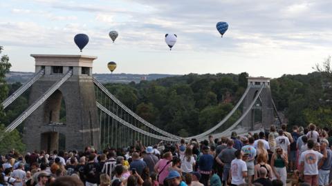 Hundreds of people stand staring at hot air ballons which are rising in the distance behind the Clifton Suspension Bridge