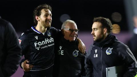 Tom Lockyer (left) smiles as he walks with his arm around the shoulders of Steve Evans (middle) at full-time of a match
