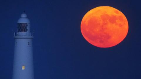 A red-orange full Moon hangs low in the night sky next to a lighthouse